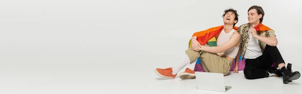 excited nonbinary person laughing near partner while sitting with rainbow flag near laptop on grey background, banner - Fotografie, Obrázek