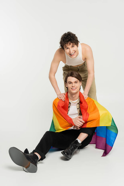 overjoyed nonbinary person smiling near partner sitting with rainbow flag on grey background - Foto, Imagem