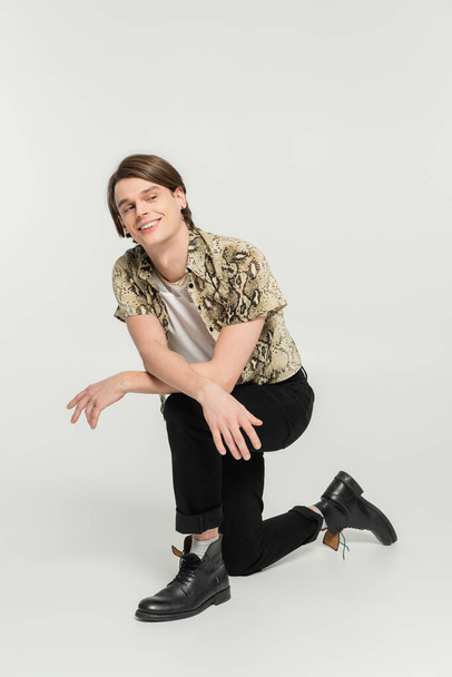 full length of cheerful nonbinary model in black pants and animal print blouse looking away on grey background - Fotó, kép