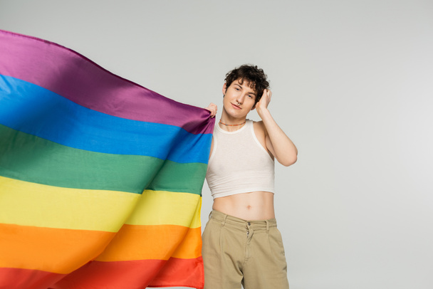  stylish bigender person in crop top and beige pants standing near rainbow flag isolated on grey - Φωτογραφία, εικόνα