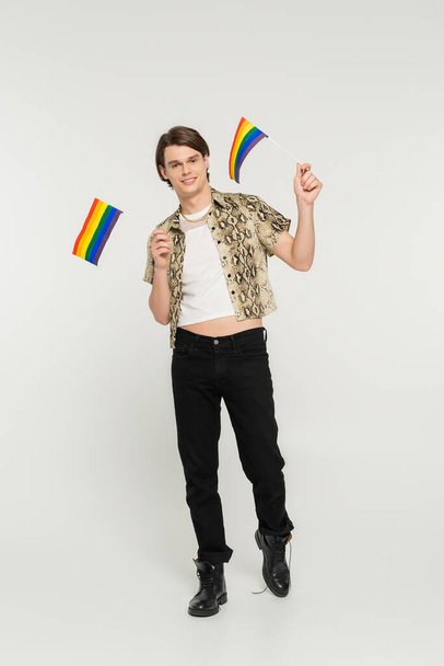 full length of positive pansexual model in black pants and snakeskin print blouse holding small lgbt flags on grey background - Foto, Imagem