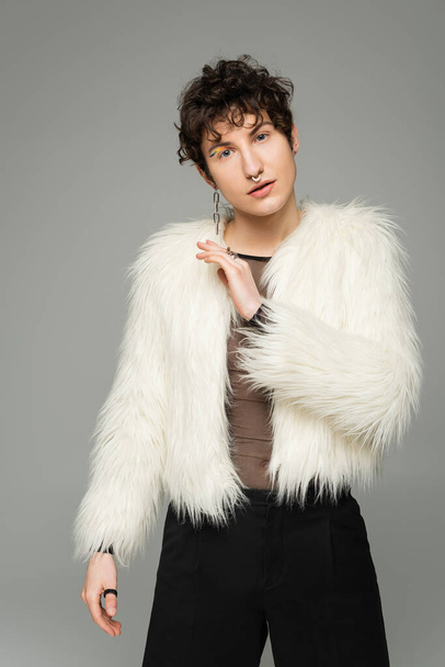 fashionable nonbinary person in white faux fur jacket looking at camera isolated on grey - Фото, изображение