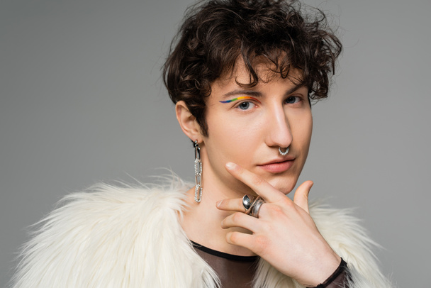 portrait of brunette nonbinary person with rainbow eye liner and silver accessories holding hand near face isolated on grey - 写真・画像