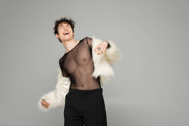 carefree nonbinary model in black transparent top and white faux fur jacket laughing with closed eyes isolated on grey - Foto, immagini