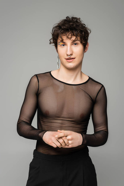 positive pansexual person in black transparent top and silver earring looking at camera isolated on grey - Fotografie, Obrázek
