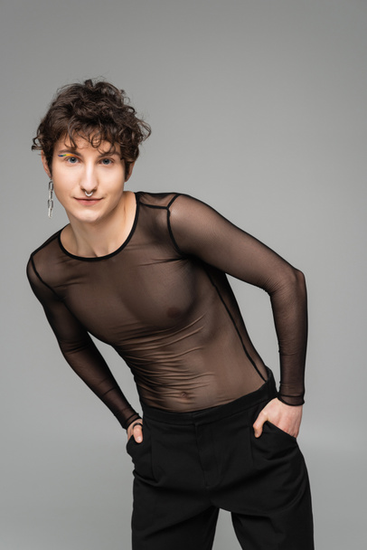 trendy bigender person in black transparent top posing with hands in pockets isolated on grey - Photo, Image