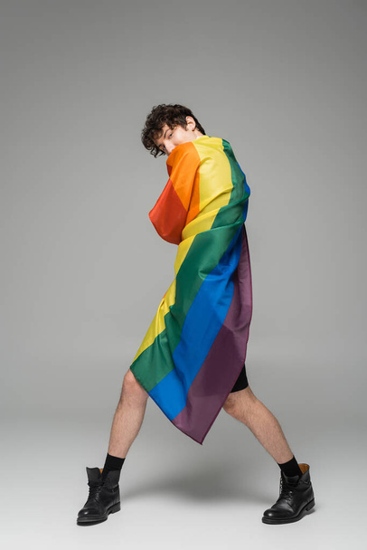 full length of nonbinary person in black boots wrapping himself in lgbt flag on grey background - Фото, изображение
