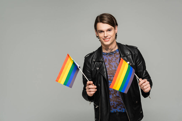 happy pangender person in black leather jacket holding small lgbt flags and smiling at camera isolated on grey - Fotó, kép