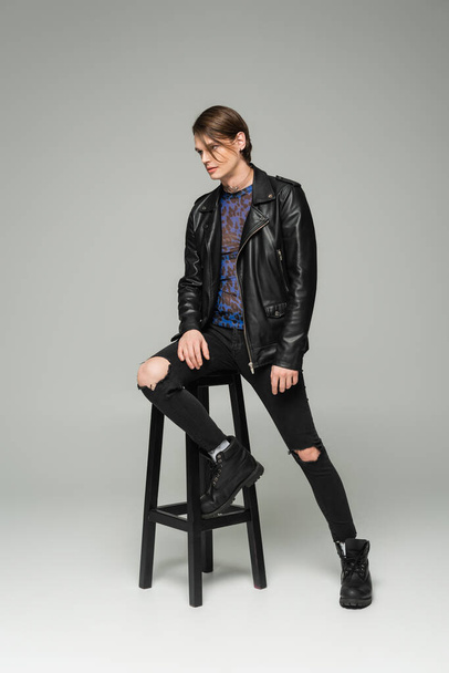 plná délka bigender modelu v černé kožené bundě a roztržené kalhoty pózující v blízkosti vysoké stoličky na šedém pozadí - Fotografie, Obrázek