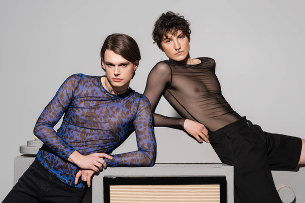 nonbinary couple in black and animal print tops leaning on huge model of photo camera on grey background - Photo, Image