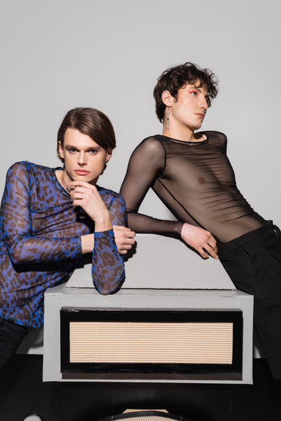 young and stylish pansexual partners posing near big model of photo camera on grey background - Photo, Image