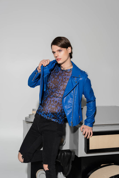 thoughtful bigender person in blue leather jacket looking away near huge model of photo camera on grey background - Foto, Bild