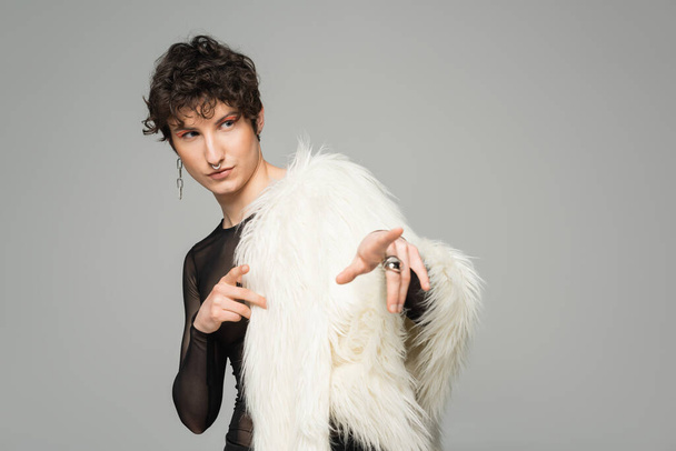 fashionable nonbinary model in white faux fur jacket pointing with fingers and looking away isolated on grey - Foto, Bild