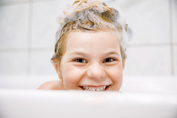 Cute child with shampoo foam and bubbles on hair taking bath. Portrait of happy smiling preschool girl health care and hygiene concept. Washes hair by herself - Фото, изображение
