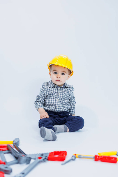 A little boy in a checkered shirt in the image of a builder. A boy in a construction helmet and plastic tools - Фото, изображение