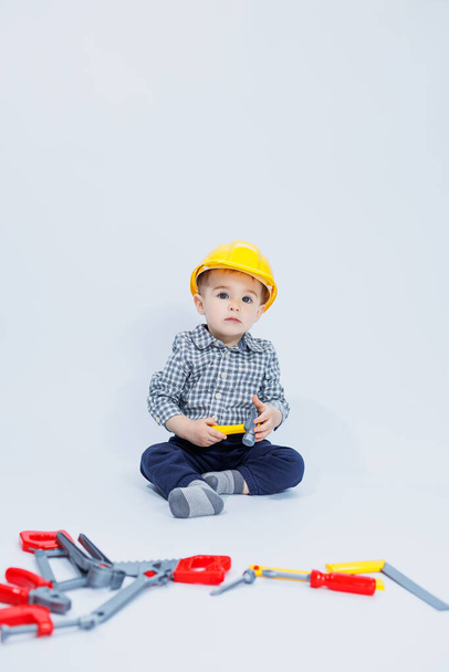 A little boy in a checkered shirt in the image of a builder. A boy in a construction helmet and plastic tools - Fotó, kép