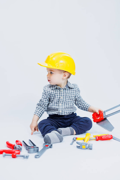 A little boy in a checkered shirt in the image of a builder. A boy in a construction helmet and plastic tools - Fotografie, Obrázek