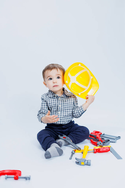 Ένα μικρό αγόρι με καρό πουκάμισο στην εικόνα ενός οικοδόμου. Ένα αγόρι με κράνος και πλαστικά εργαλεία. - Φωτογραφία, εικόνα