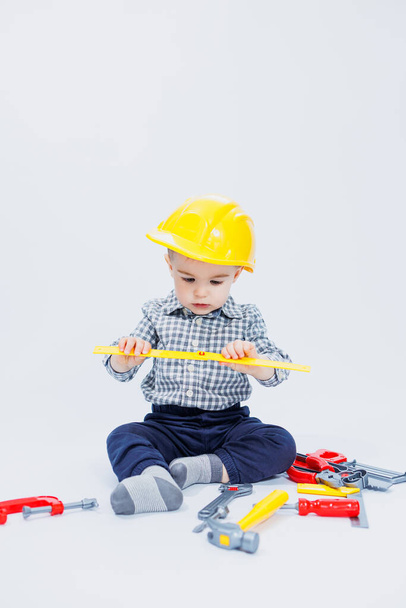 Een kleine jongen in een shirt met een helm en een speelgoedgereedschap. Witte achtergrond. Bouw speelgoedgereedschap voor kinderen - Foto, afbeelding