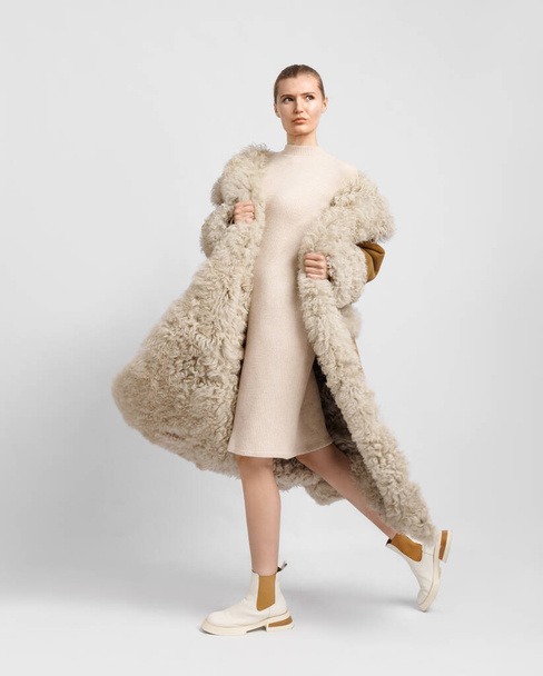 A young stylish girl in a luxurious long brown sheepskin coat with a fur hood and cuffs poses beautifully on a white background. Fashionable, stylish, comfortable outerwear. - Fotó, kép