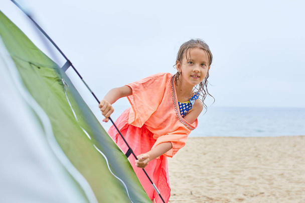 Uma criança menina feliz olha para fora de uma tenda na praia, férias em família de verão, fuga local, estilo de vida ativo saudável - Foto, Imagem