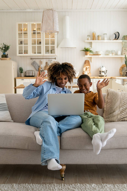 Heureuse mère afro-américaine joyeuse et son fils assis sur le canapé avec un ordinateur portable en agitant les mains et en regardant la caméra Web, avoir un appel vidéo avec la famille. Sourire maman et enfant garçon parler avec des parents en ligne - Photo, image