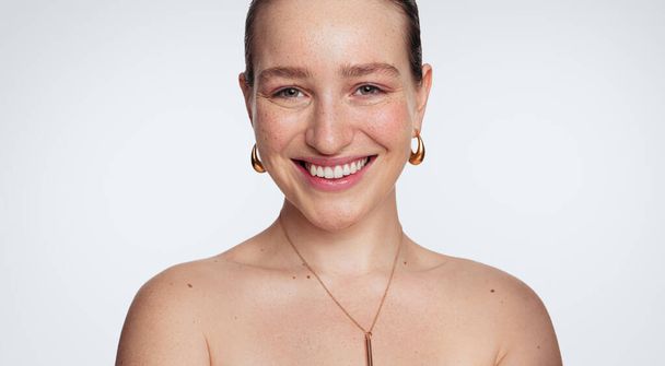 Crop confident young female model with perfect glowing skin and bare shoulders smiling happily and looking at camera against white background in light studio - Photo, image