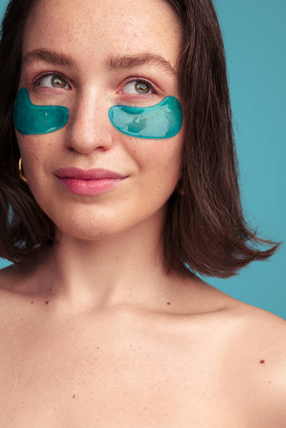 Crop young female millennial with dark hair freckles and collagen eye patches looking away during skincare daily procedure in blue studio - Foto, Bild
