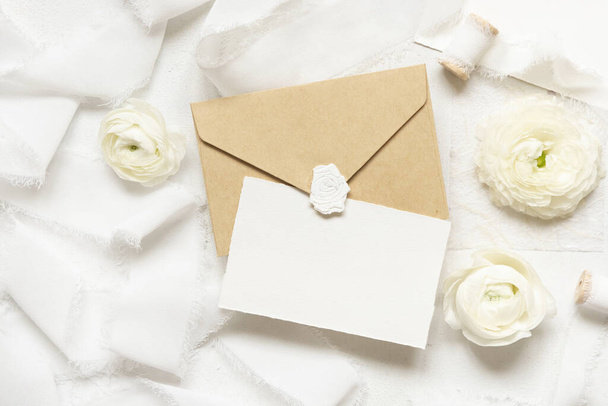 Blank card and envelope near cream roses and white silk ribbons top view,  wedding mockup. Romantic scene with vertical card and pastel flowers flat lay. Valentines, Spring or Mothers day concept - Fotó, kép