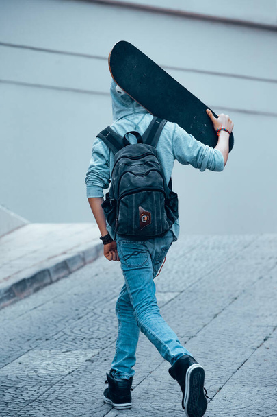 Backside of a teenager with backpack walking with skateboard in hands. Active boy outdoors. Happy time of young people. Sportive lifestyle. - Fotoğraf, Görsel