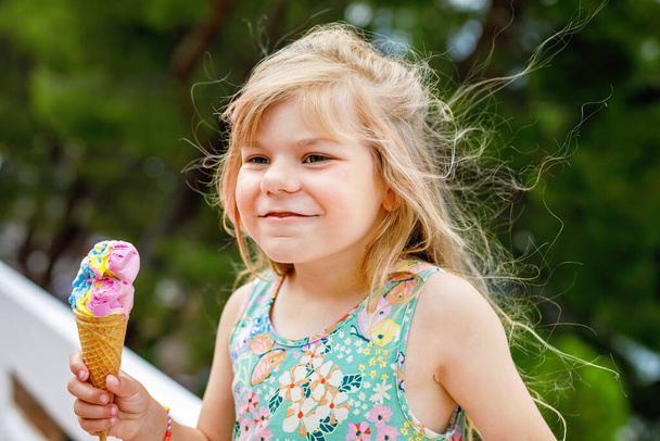 Boldog óvodás lány eszik színes jégkrémet gofris tölcsérben a napsütéses nyári napon. A kisgyerek jégkrém desszertet eszik. Édes étel meleg nyári napokon. Élénk fény, színes fagylalt. - Fotó, kép