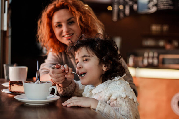 Όμορφη νεαρή κοκκινομάλλα μητέρα με χαριτωμένο σγουρά μαλλιά κόρη κάθονται σε ζεστό καφέ και πίνουν ζεστό σχολείο. Ημέρα μητέρων. - Φωτογραφία, εικόνα