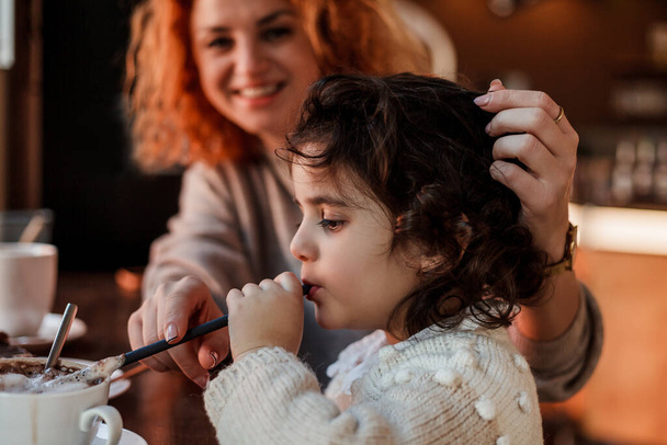 Beautiful young red-haired mother with cute curly-haired daughter are sitting in cozy cafe and drinking hot school. Mothers Day. - Photo, Image