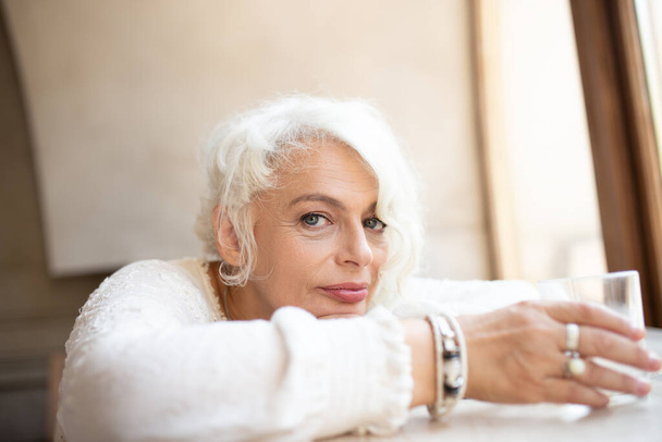 Портрет расслабленной мечтательной женщины в возрасте с белыми волнистыми волосами, сидящей у окна, опирающейся головой на руки и держащей стакан воды, смотрящей в камеру с выражением спокойствия и спокойствия - Фото, изображение