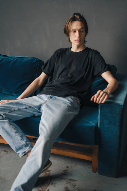 Portré egy vidám fiatalemberről, aki alkalmi fekete pólóban ül otthon a kanapén. Modern életmód. Emberek életmód portréja. Hangulatos apartman. - Fotó, kép