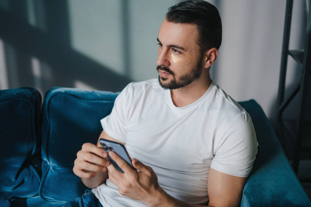 Young serious man using modern smartphone device while sitting on sofa at home, typing an sms message at social network, technology concept. Technology - Fotó, kép