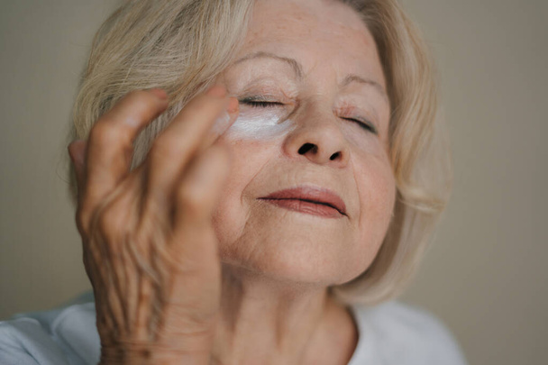 Großaufnahme Porträt einer Seniorin, die unter der Augencreme aufträgt, wodurch ihre Haut sof wird. Faltenfrei mit natürlicher Anti-Aging-Kosmetik. Produkt für Schönheit - Foto, Bild