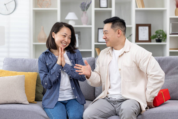 Happy asian family man and woman together at home sitting on sofa in living room, husband giving gift to wife in celebration, couple hugging and happy. - Фото, зображення