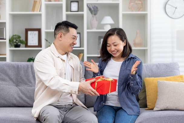 Happy asian family man and woman together at home sitting on sofa in living room, husband giving gift to wife in celebration, couple hugging and happy. - Photo, image