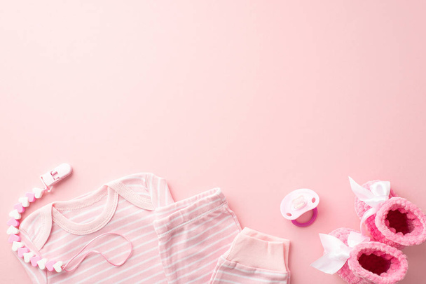 Baby accessories concept. Top view photo of pink infant clothes shirt pants dummy teether chain and knitted booties on isolated pastel pink background with empty space - Photo, Image
