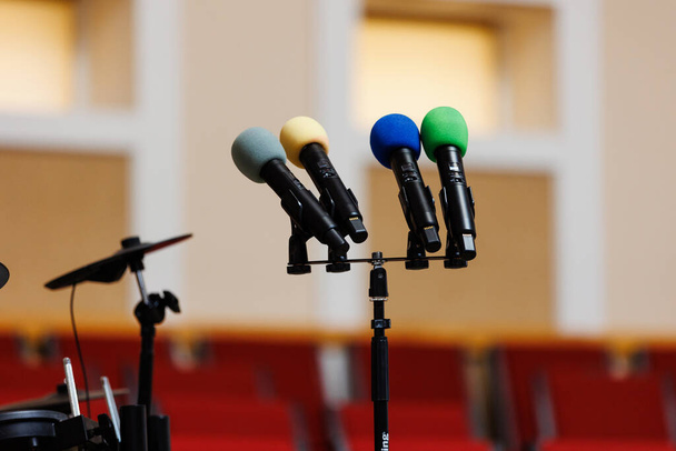 Cztery kolorowe mikrofony na stojaku na podium i wiele czerwonych krzeseł rozmytych w tle. Koncepcja komunikacji, wydarzenia, seminarium, media, konferencja prasowa, performance, polityka, firma - Zdjęcie, obraz