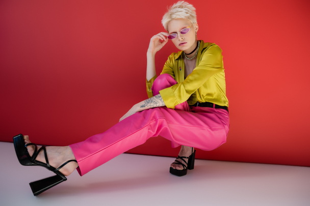 full length of tattooed albino model in trendy heeled sandals and sunglasses posing on carmine pink  - Фото, зображення