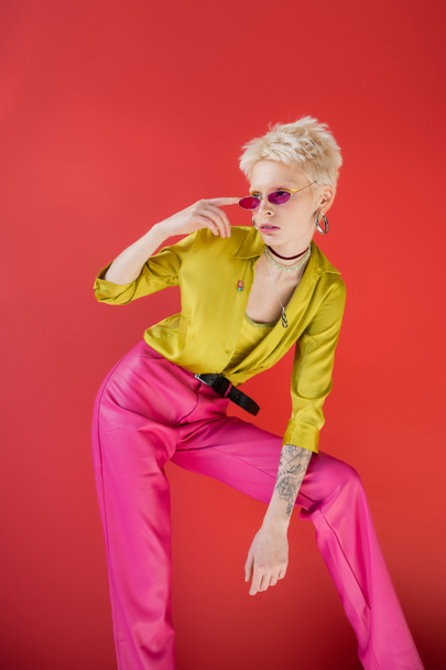 albino model with tattoo posing in stylish blouse and adjusting trendy sunglasses on carmine pink  - Foto, Imagem