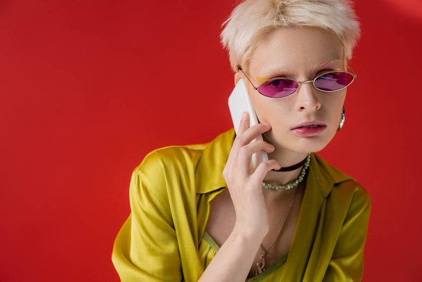 albino woman in trendy sunglasses talking on smartphone on carmine pink background  - Photo, Image