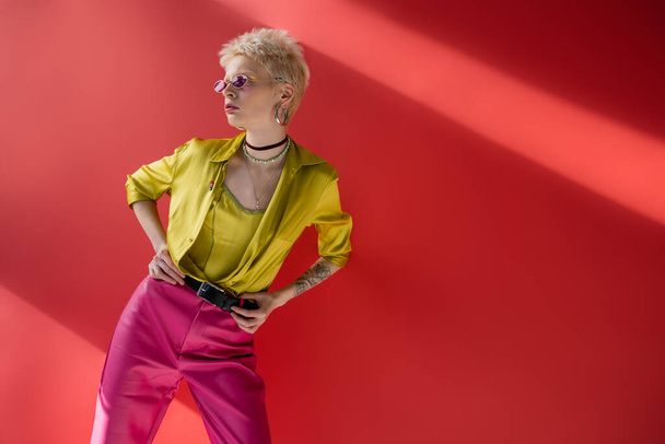 young albino model with tattoo on hand posing in stylish blouse and trendy sunglasses on carmine pink background  - Photo, Image