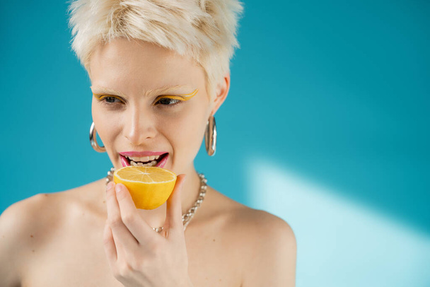 blonde albino woman with bare shoulders biting sour lemon half on blue background  - Photo, Image