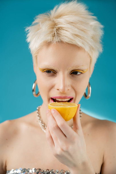 blonde albino model with bare shoulders biting sour lemon half on blue background  - Фото, изображение