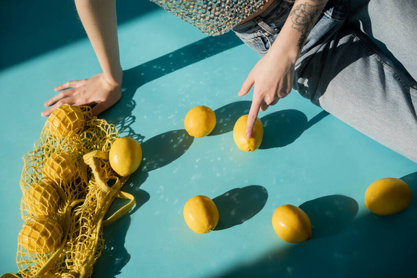 cropped view of tattooed woman in shiny top with sequins and jeans sitting near string bag with ripe lemons on blue  - Photo, Image