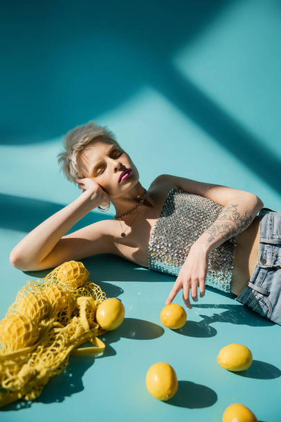 tattooed albino model in shiny top with sequins and jeans posing near ripe lemons on blue  - Photo, Image