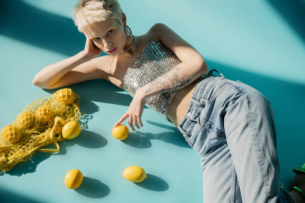 shora pohled na tetovanou albínskou ženu nahoře s flitry a džínami ležícími poblíž zralých citronů na modré  - Fotografie, Obrázek